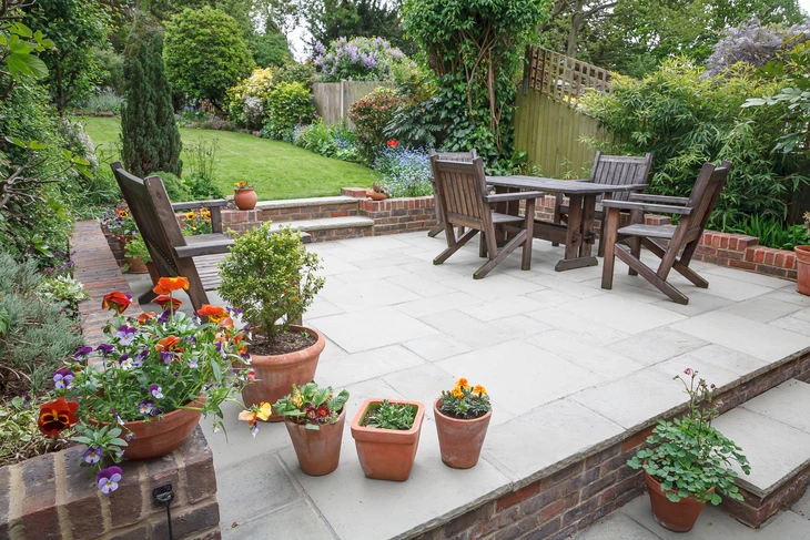 Terrasse extérieure aménagée avec salon de jardin en bois, jardin paysager en arrière-plan, bordures végétalisées et pots de fleurs colorées