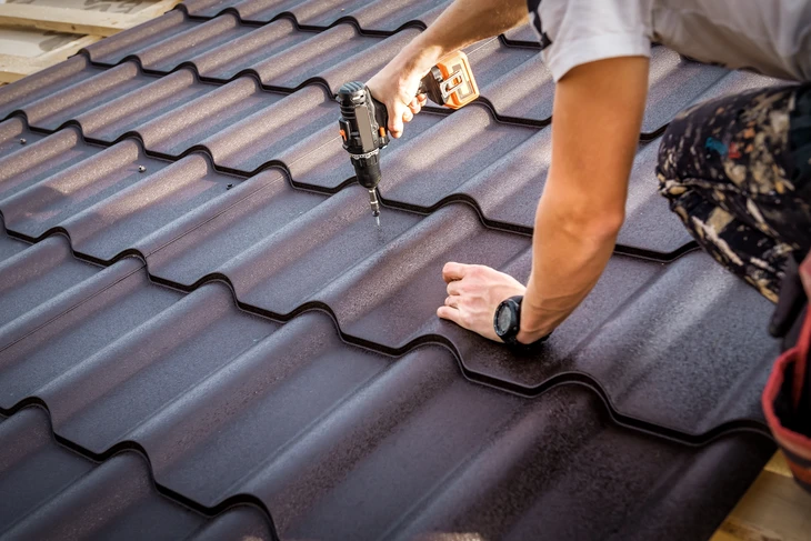 Ouvrier sur toiture en tuiles métalliques ondulées grises, utilisant une perceuse-visseuse sans fil pour fixer les éléments de couverture