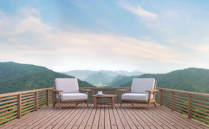 Terrasse en bois avec deux fauteuils modernes à coussins gris et table basse, surplombant une vallée montagneuse boisée