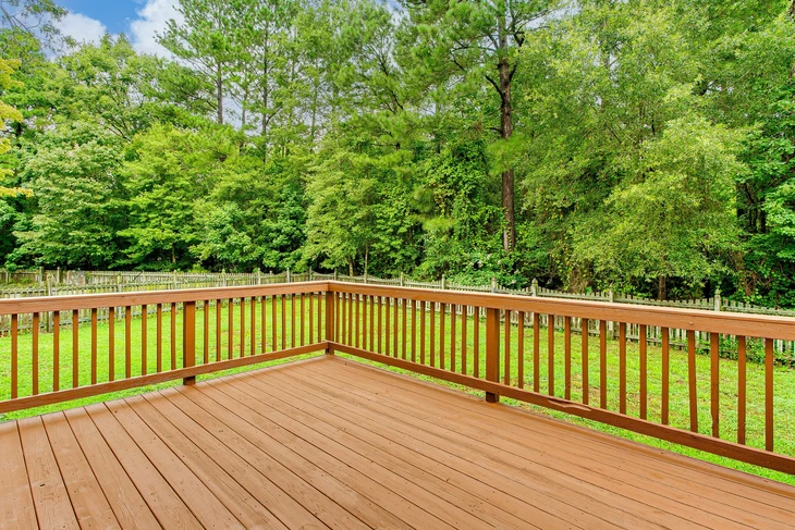 Terrasse extérieure en bois avec garde-corps à barreaux verticaux donnant sur jardin verdoyant et forêt dense d'arbres feuillus