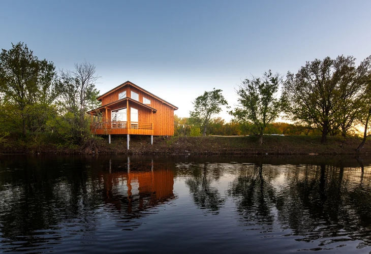 Maison contemporaine en bois sur pilotis au bord d'un étang, entourée d'arbres au coucher du soleil