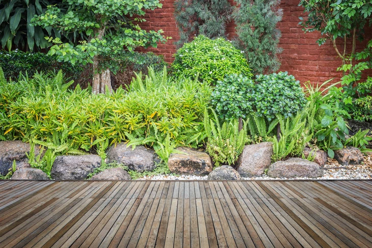 Jardin paysager extérieur avec terrasse en bois composite, bordure de pierres naturelles et plantation mixte de fougères, arbustes persistants et conifères nains contre mur de briques rouges