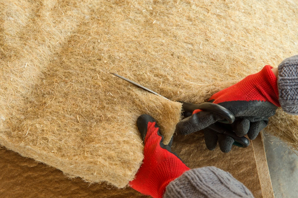 Travailleur en gants de protection rouge et noir découpant de l'isolant en fibre de coco avec un cutter sur chantier de construction