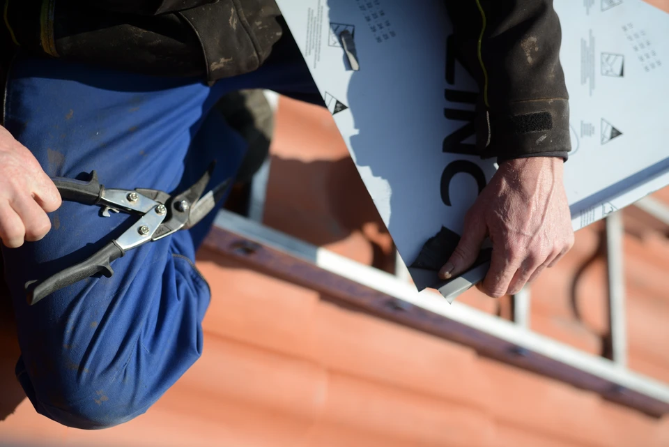 Photographie d'un chantier de construction bois en extérieur montrant deux ou trois générations d'artisans (un homme mature expérimenté, un artisan d'âge moyen et un jeune apprenti) travaillant ensemble sur la pose d'une charpente ou d'une couverture. Les ouvriers portent des équipements de protection appropriés et travaillent de manière coordonnée sur la structure d'une maison, illustrant la transmission du savoir-faire et la collaboration générationnelle. L'ambiance doit être sérieuse et professionnelle, montrant la précision et l'expertise du métier artisanal.